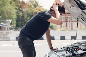 the man stands in front of the car with the bonnet open, leaning over the engine and scratching his head thoughtfully